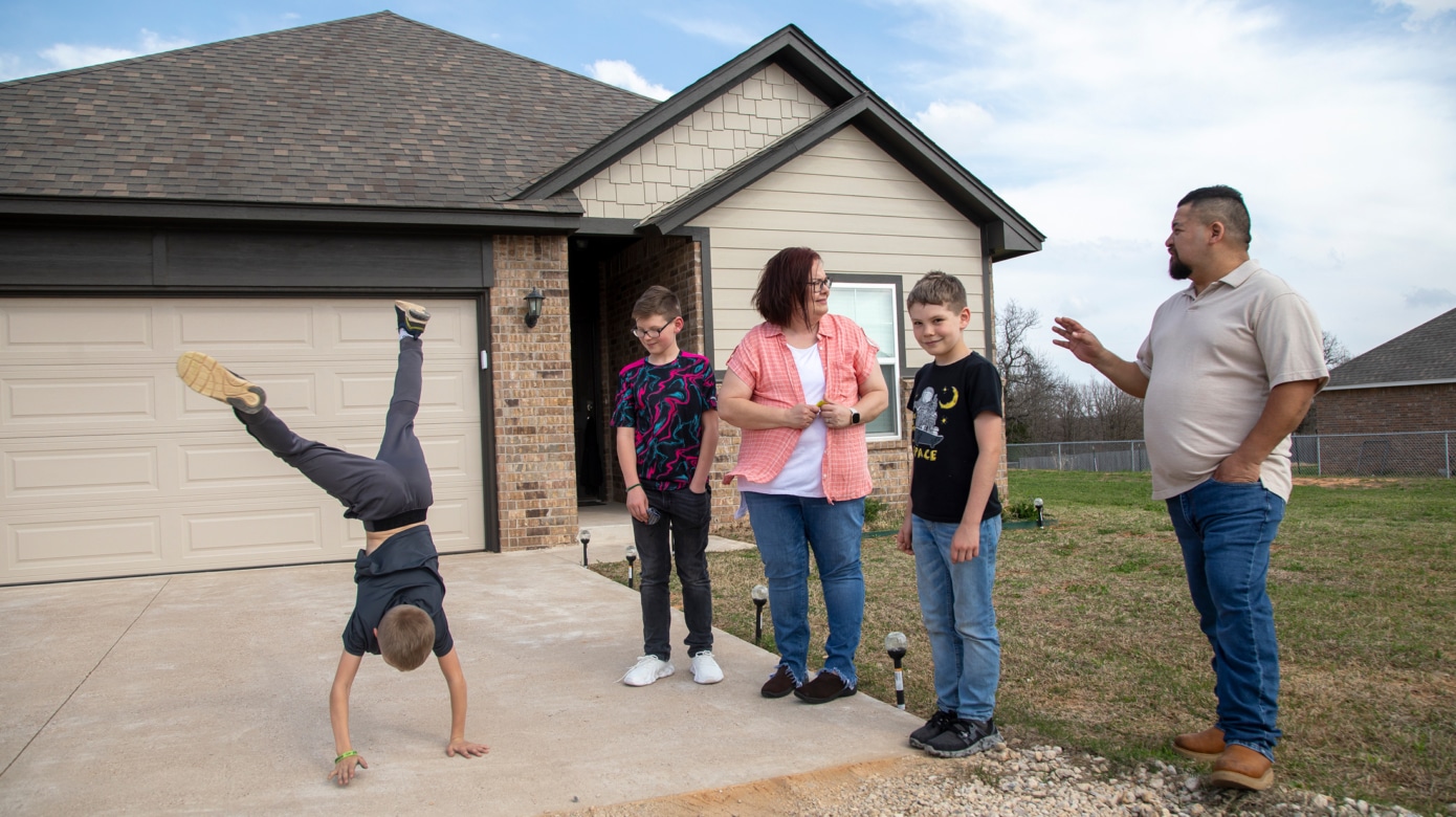A family of five gathers in front of their new home. The parents and two kids stand candidly, talking, and one child performs a cartwheel across the driveway.