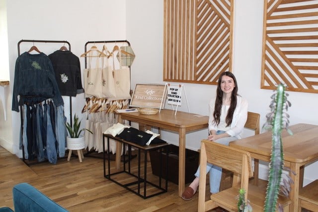 Tribal member Hannah Muller sits in a clean, neutral toned room with racks of embroidered clothing she makes for her business, Kind Collections.