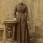 Full body sepia tone portrait of Julia Anderson. She stands beside a stone memorial and wears a long dress with buttons down the front.