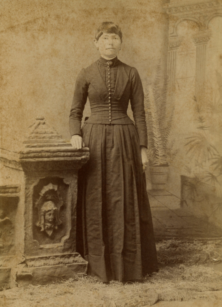 Full body sepia tone portrait of Julia Anderson. She stands beside a stone memorial and wears a long dress with buttons down the front.