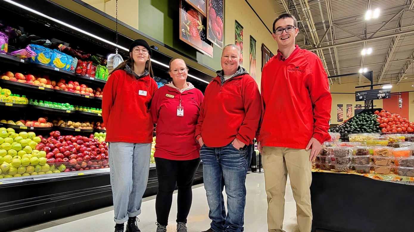 Four FireLake Foods employees wearing red FireLake polos stand in the produce department at FireLake Discount Foods. Apples, tomatoes, avocados and more are visible over their shoulders.
