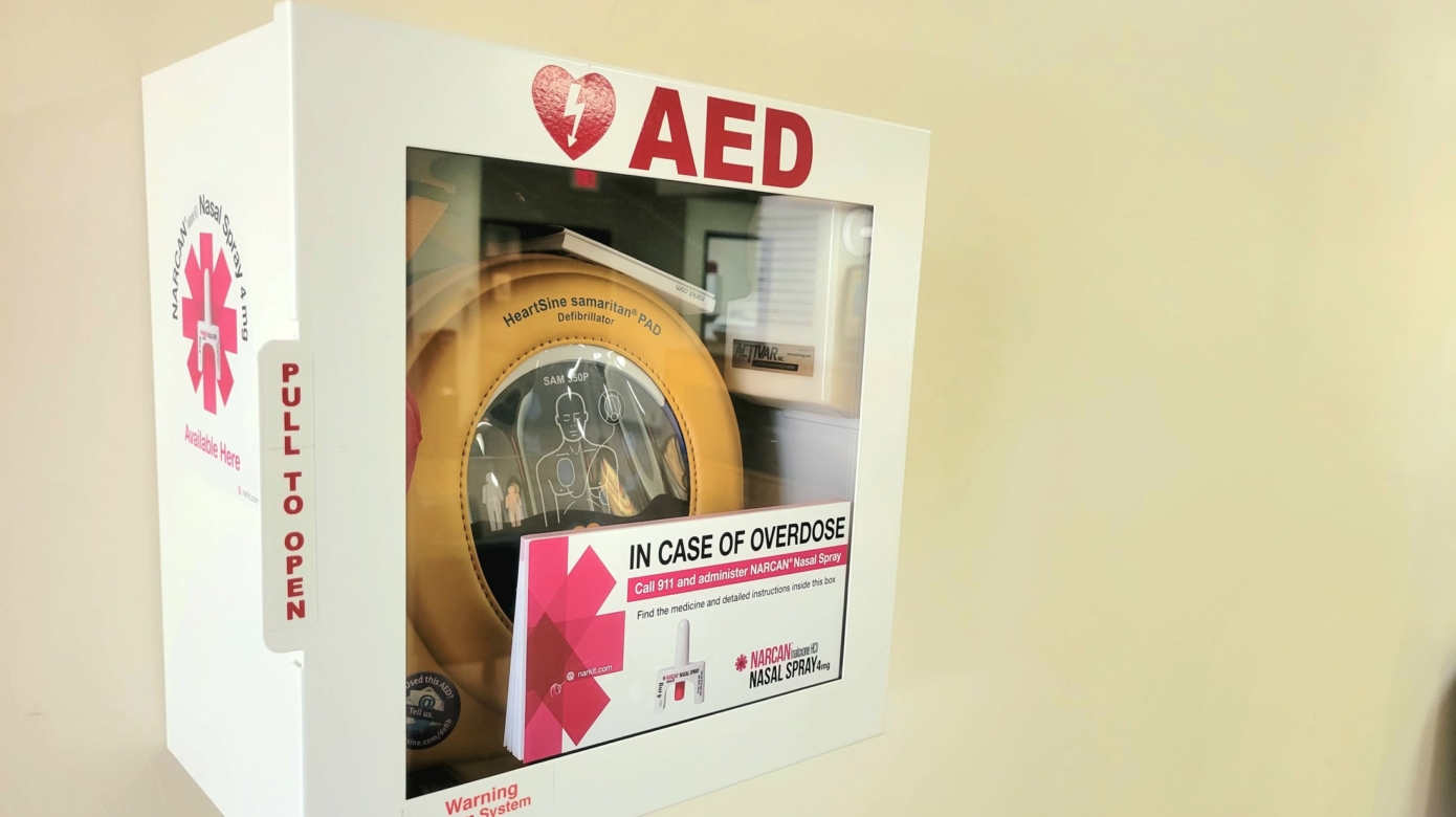 AED box with bright pink signage indicating NARCAN kits and instructions included in the box.