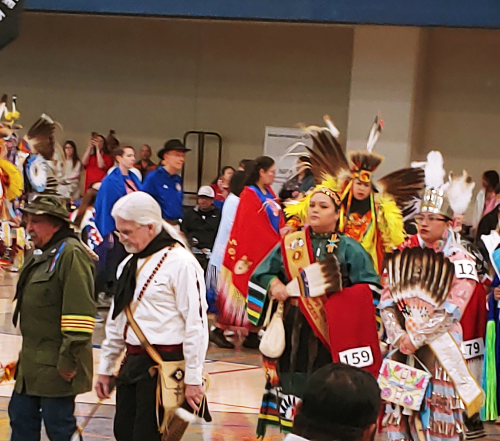 A crowd of people in colorful regalia enter an indoor dance arena.