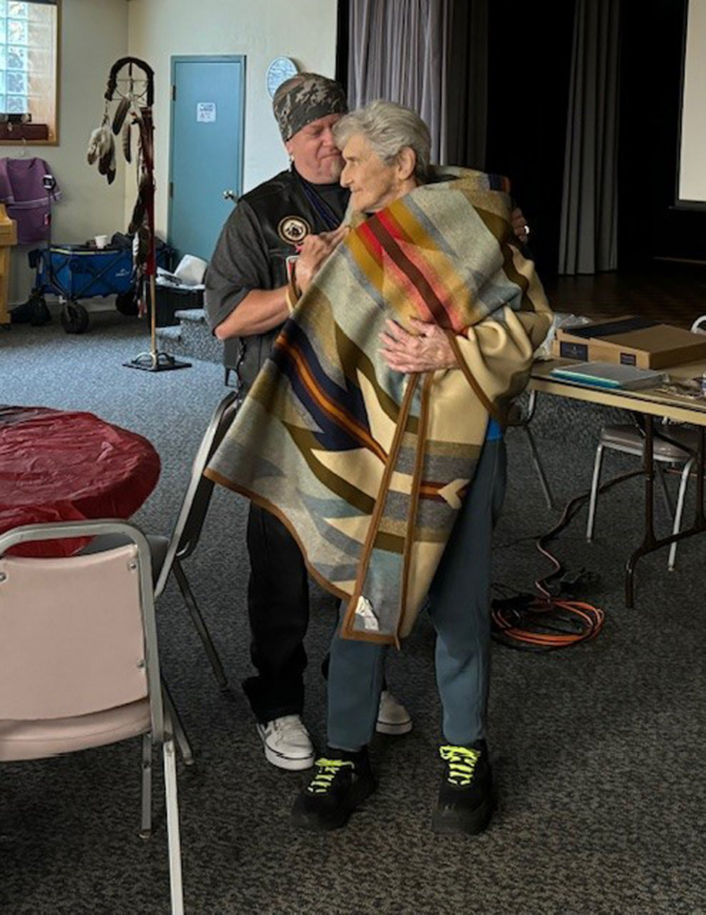 A CPN Elder is wrapped in a Pendleton blanket by another Tribal member.