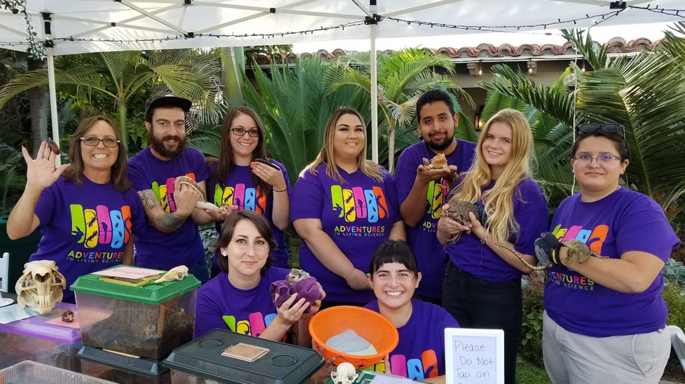 Several people wearing purple Adventures in Living Science t-shirts hold small animals at an education event.