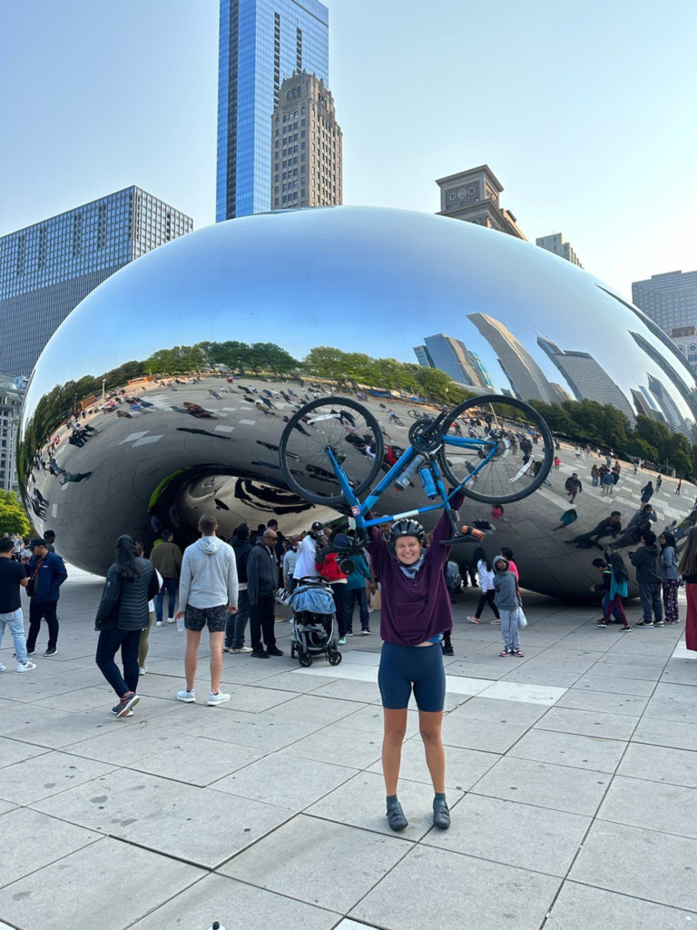Athlete Alicia Rusthoven, wearing biker shorts and a purple sweatshirt, holds her blue bicycle over her head in front of the iconic Cloud Gate statue (the Bean) in Chicago, the skyline and fellow tourists reflected in its surface around her.