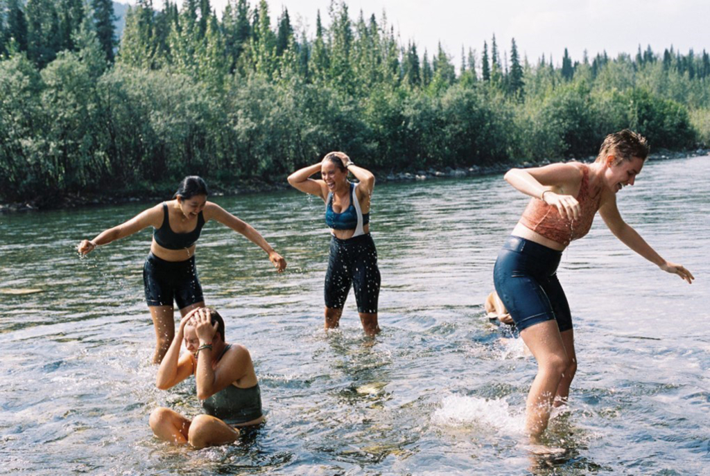 Four athletes splash in a stream with evergreen forests in the background.