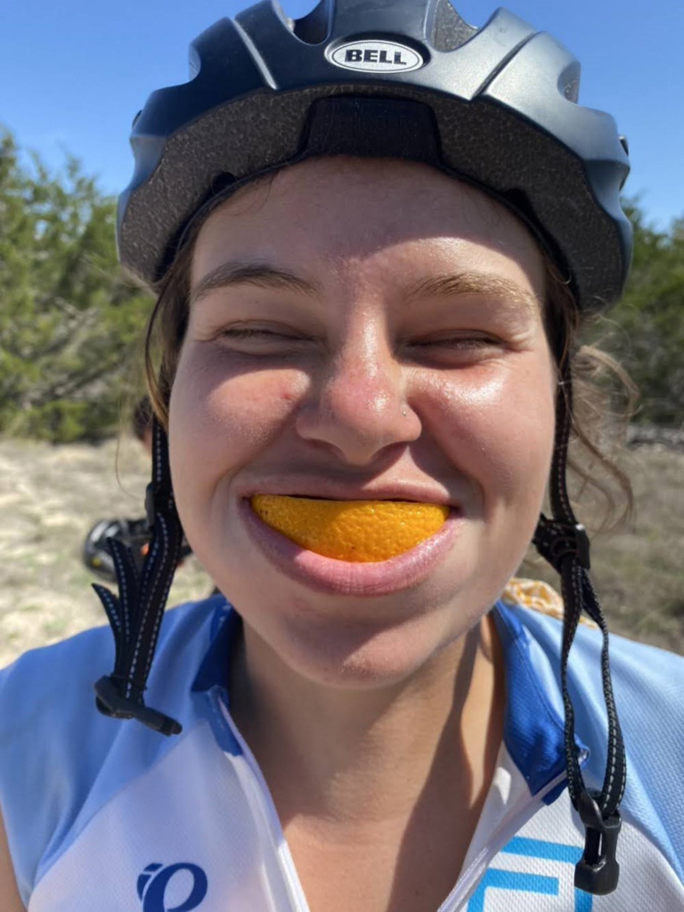 Alicia Rusthoven, wearing a bicycling suit and helmet, smiles with an orange slice in her mouth.