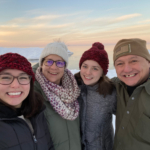 A selfie with four family members in a snowy landscape with a pastel sunset behind them. Jozie and her sister, Tessa, wear maroon beanies. Her mother Tondra, between the two sisters, wears a white hat, green coat, and purple scarf. Her father, Ed, is at the far right of the photograph and wears a khaki beanie with an American flag. All four smile from ear to ear.