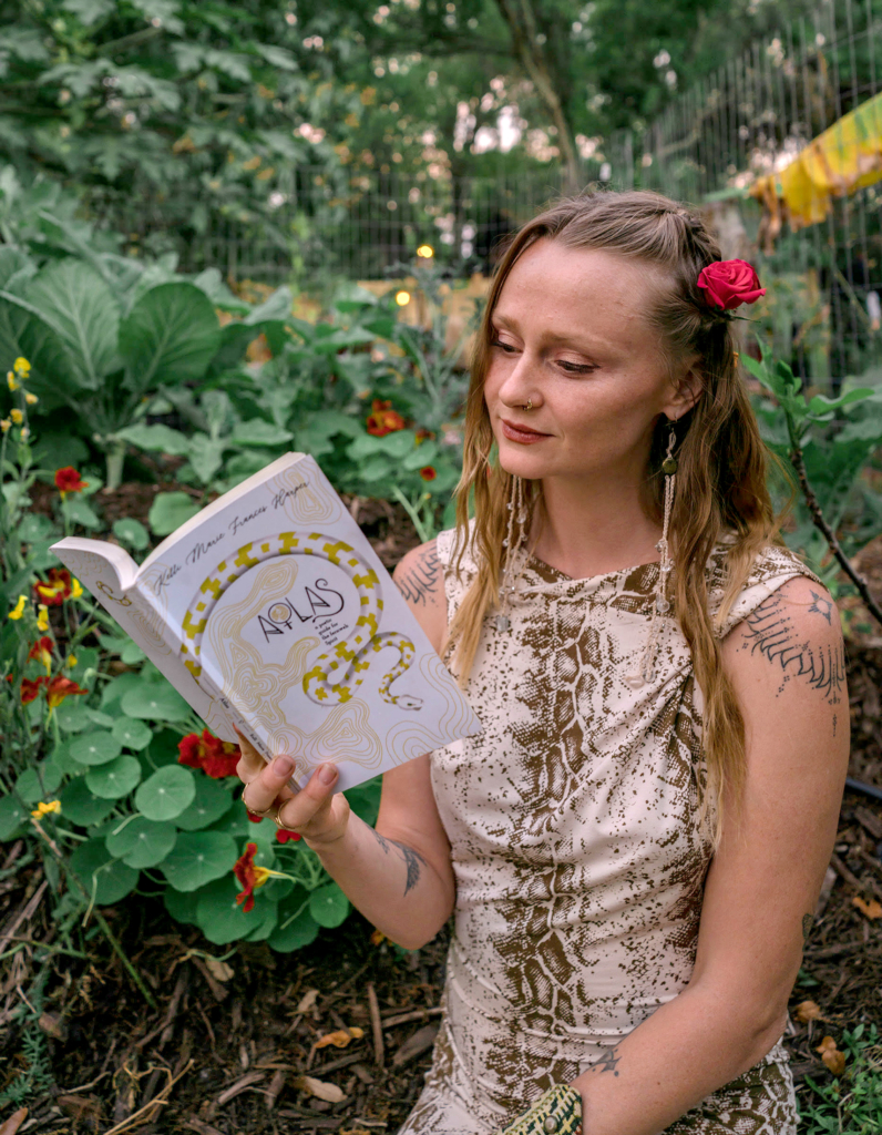 Poet Kelli Harper stands in a garden reading from her poetry book, Atlas. She has a pink flower in her blonde hair, which falls over her shoulders.