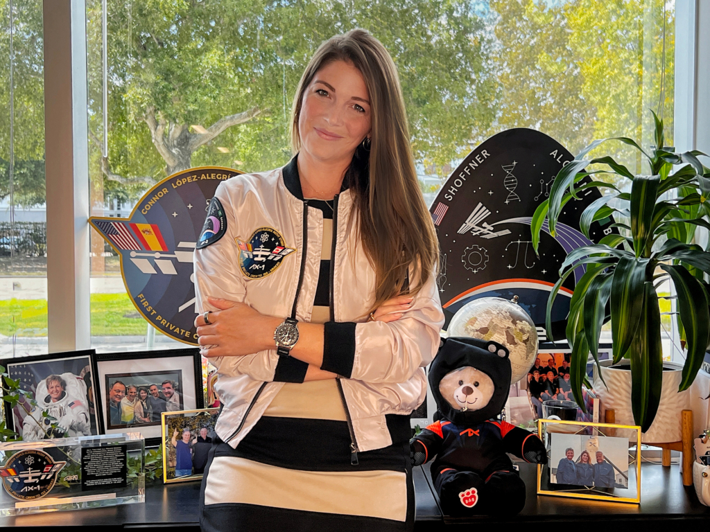 Audre Kiefer, in a white jacket with Axiom patches on it, stands in front of a window with photos and memorabilia of her accomplishments with Axiom Space.