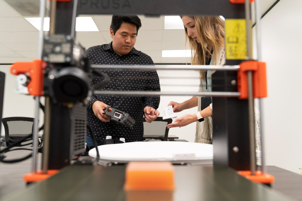 Audrey Kiefer and a colleague are visible through the orange and black apparatus of a machine as they work on the AxEMU spacesuit team together.