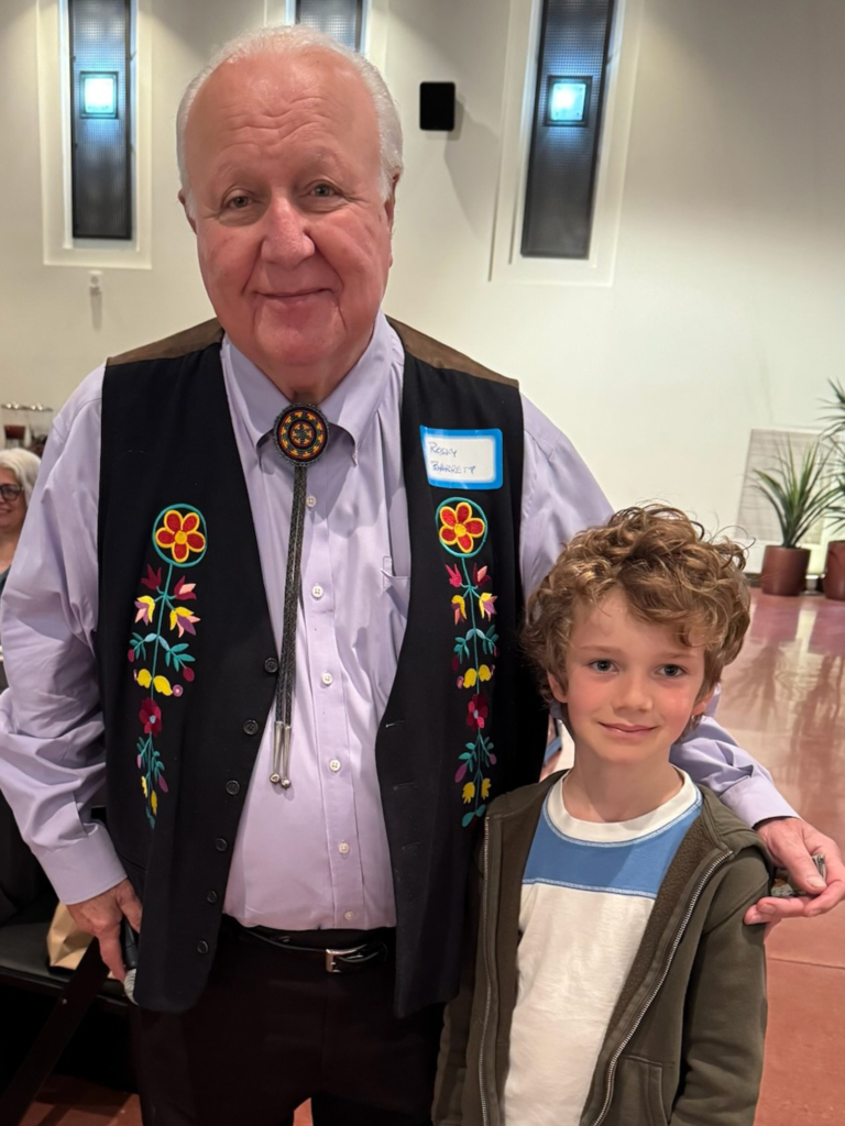 Tribal Chairman John "Rocky" Barrett stands with a young Tribal member with curly hair wearing a blue and white t-shirt and zip-up jacket.