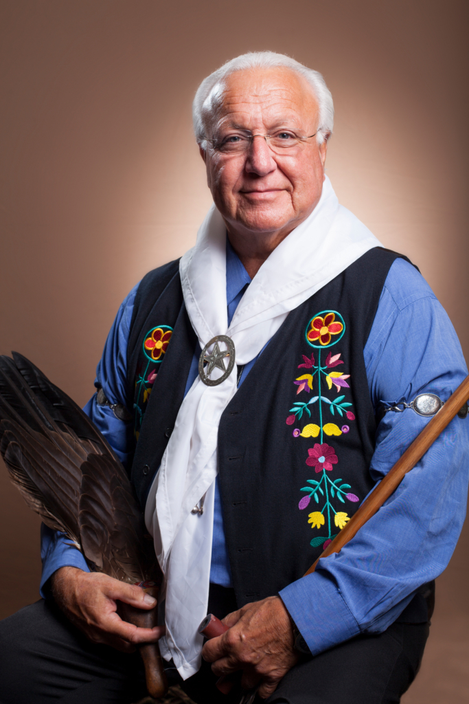 Portrait of CPN Tribal Chairman John "Rocky" Barrett. Barrett wears a blue shirt and black vest with woodland floral patterns, and carries a cane and an Eagle fan.