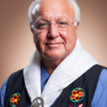 Headshot of CPN Tribal Chairman John "Rocky" Barrett, wearing a blue shirt, black vest with woodlands floral patterns, and a white scarf.