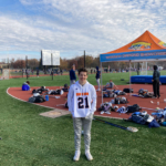 Chase Peltier, wearing a white hoodie and khaki pants, stands near the edge of a lacrosse field and poses for a photograph.