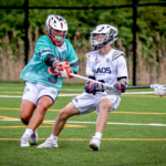 Chase Peltier, in a white lacrosse uniform, and a player in a teal jersey are photographed in action mid-game.