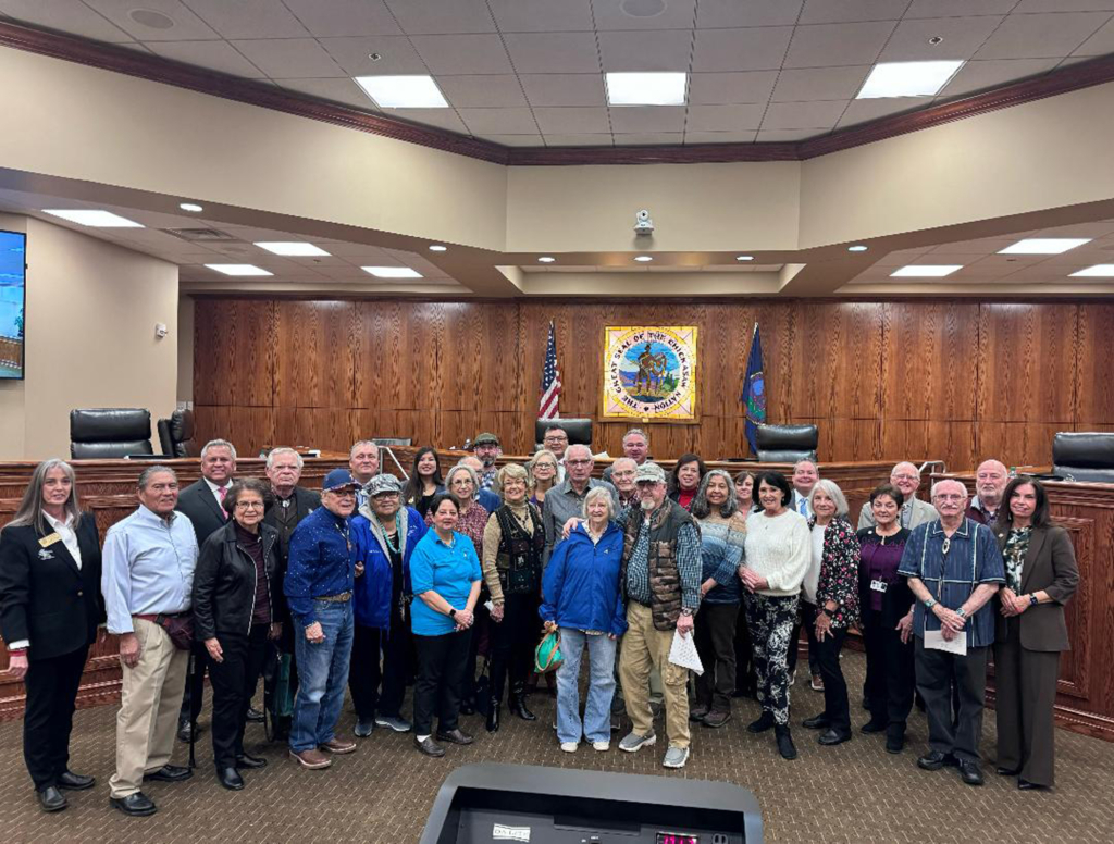 Group photo in a wood-paneled legislative room. The seal of the Chickasaw Nation is on the back wall.