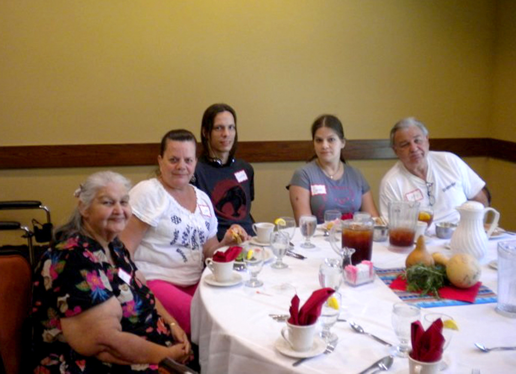 A group of five sit at a round table with a white table cloth and a tea spread.
