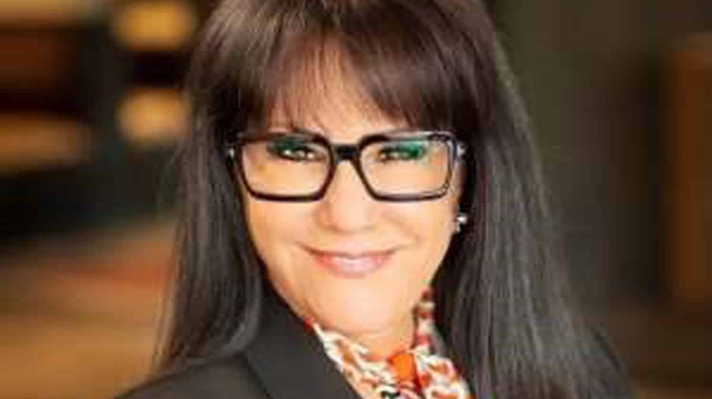 Headshot of Crissy Rumford in a black blazer and patterned blouse in warm colors. Her black glasses pop on her face, and her smile is energetic and bright.