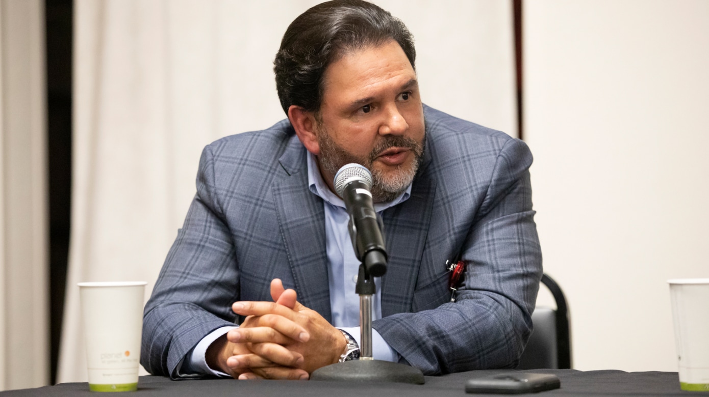 CPNHS Behavioral Health Psychologist Dr. Julio Rojas speaks into a microphone at the panelist table during the recent awareness event. He wears a grey suit jacket and blue shirt.