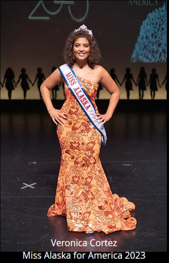 A young person wearing a peach one-shoulder gown covered in coral sequined flowers stands on a marley stage. She wears a tiara in her dark curly hair, and a sash that reads "Miss Alaska."