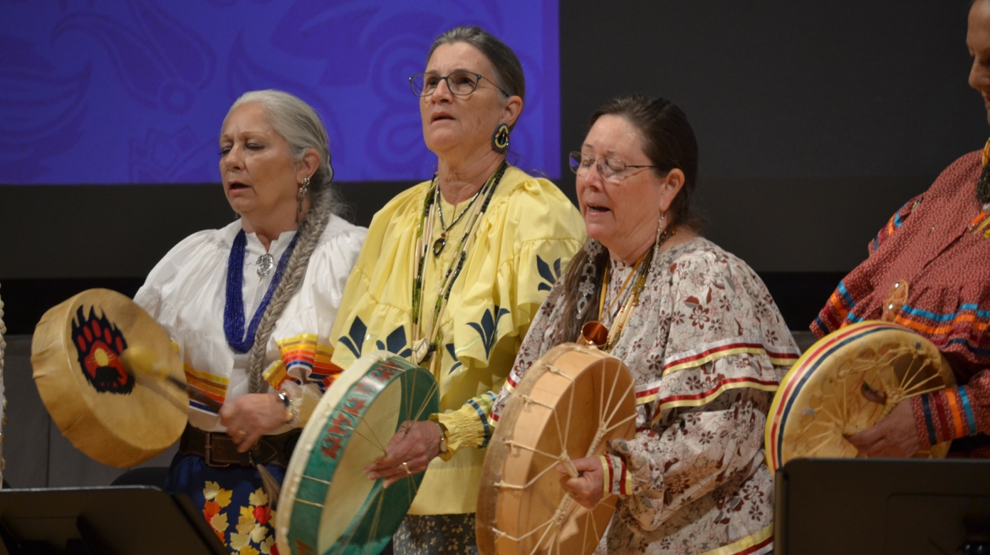 Three members of De'Wegen Kwek, Citizen Potawatomi Nation's women's drumming group, sing and drum. They wear ribbon shirts and skirts of warm colors and floral patterns, and stand behind several music stands.