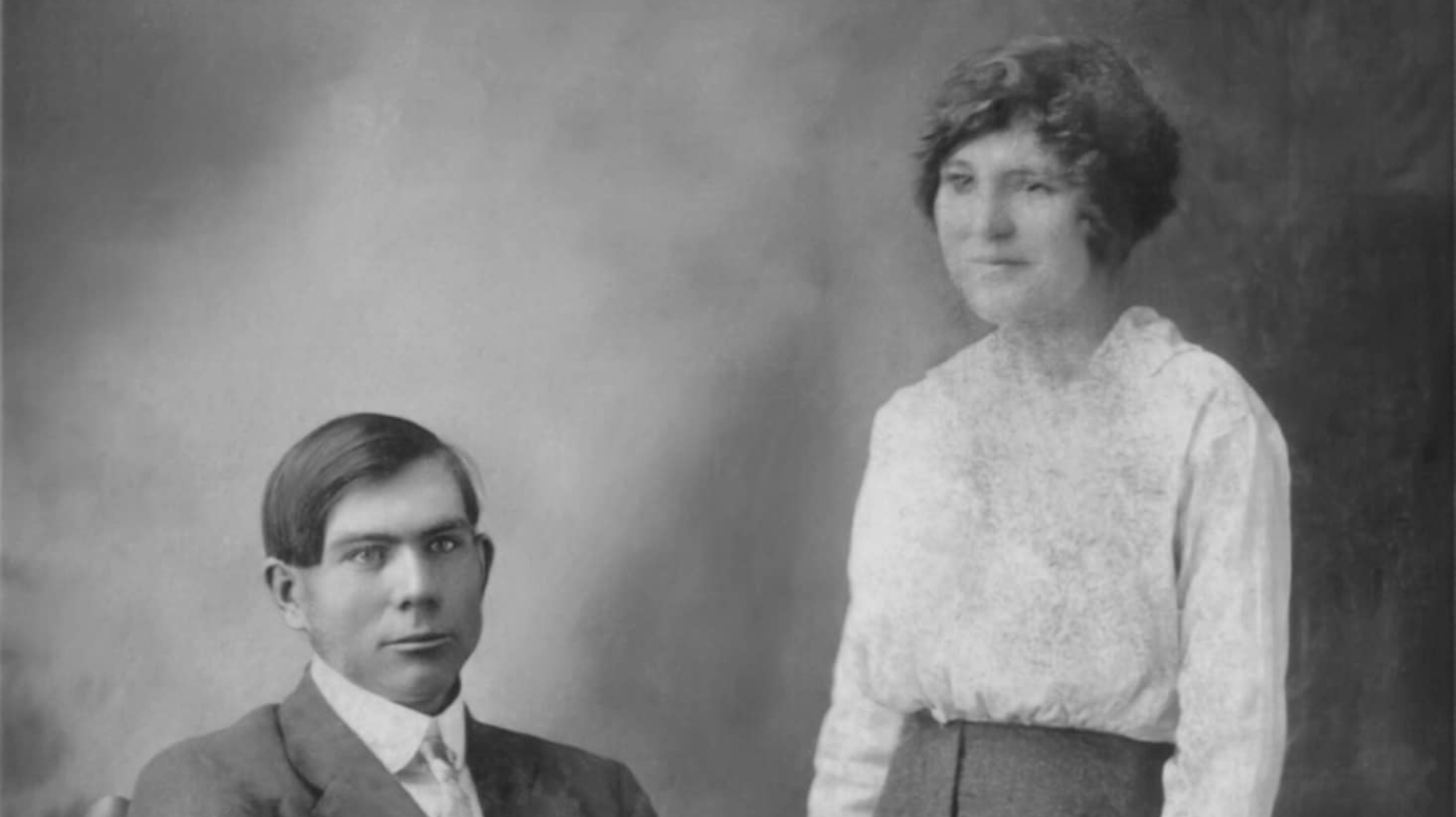 A young man, seated in a chair adorned with scrolls. A young woman in a white blouse and long skirt stands next to him.