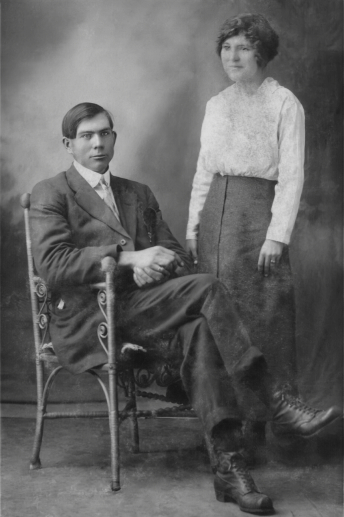 A young man, seated in a chair adorned with scrolls. A young woman in a white blouse and long skirt stands next to him.