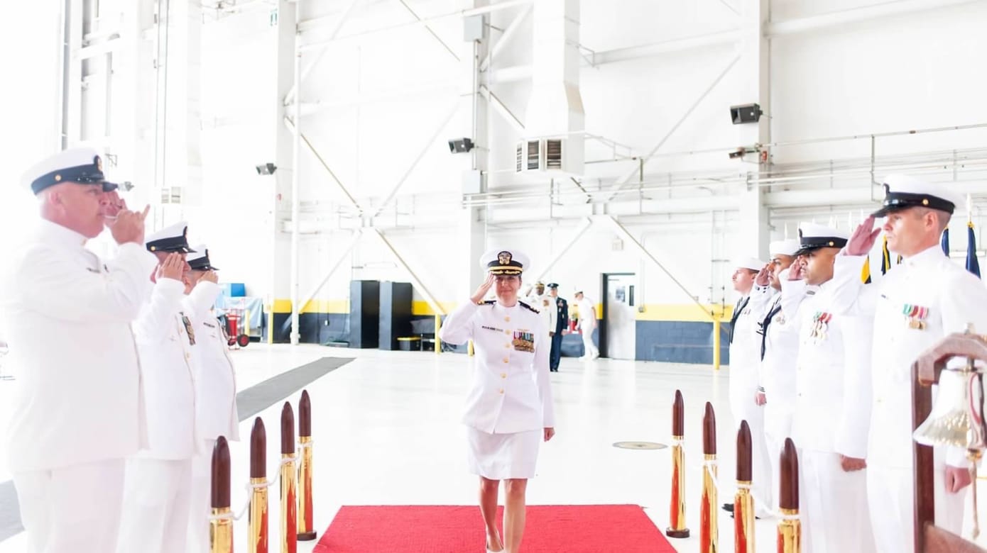 Commander Desrochers salutes as she walks towards the camera along a red carpet lined with Naval officers in dress whites on each side.