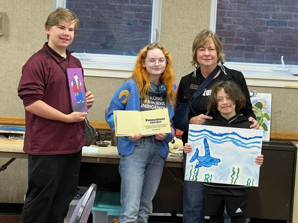 Photo of three young artists holding their painted and printed artworks and standing next to Eva Marie Carney.