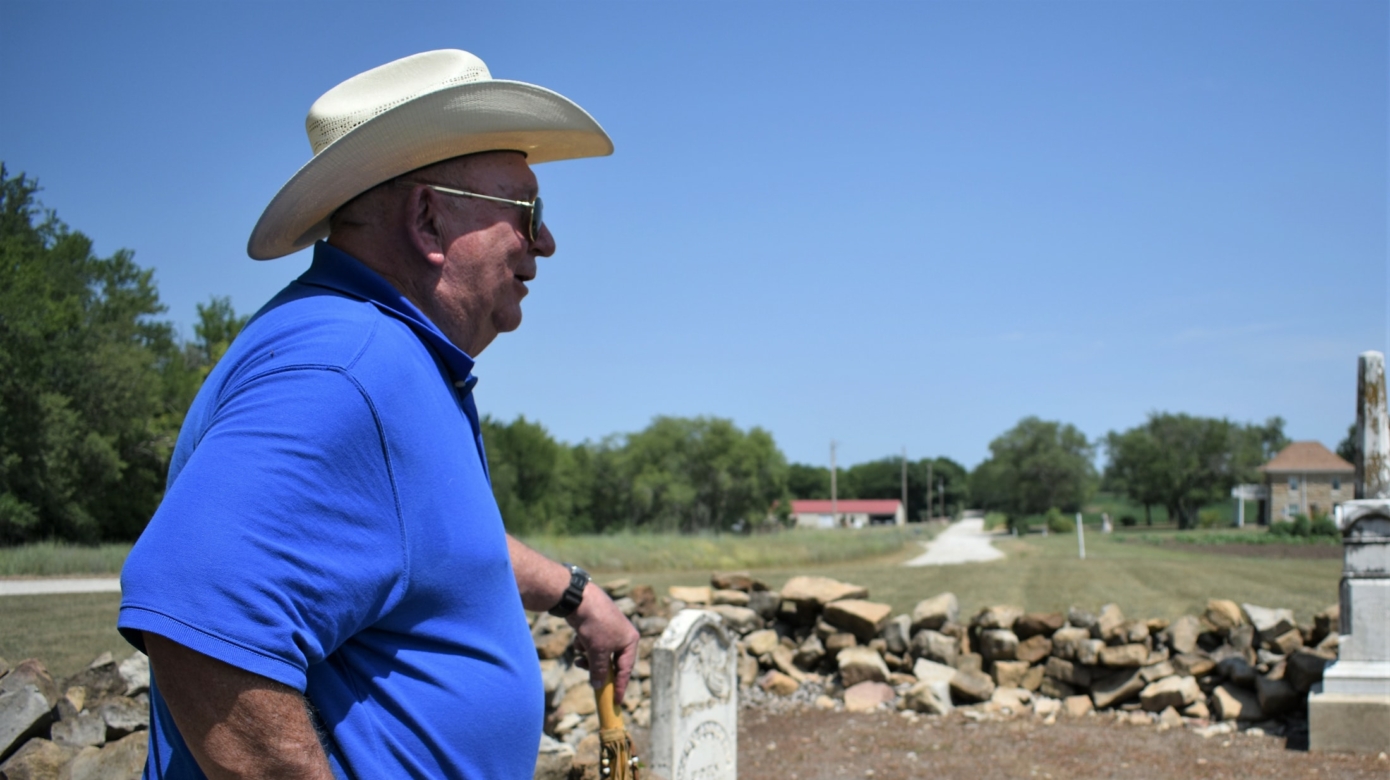 Citizen Potawatomi Nation District 4 legislator Jon Boursaw stands leaning on a staff looking over a ring of rocks at the Uniontown Cemetery during the 2018 Potawatomi Gathering of Nations. Boursaw wears a bright blue tshirt and jeans, and a light western style hat.