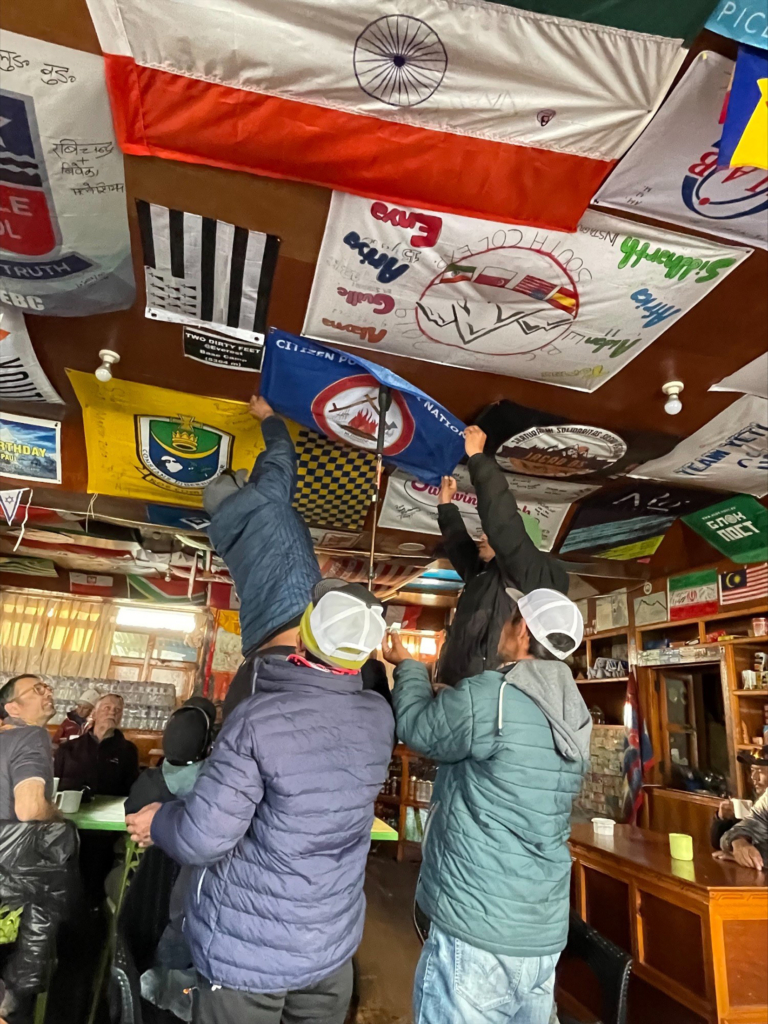 Several people in puffer jackets work together to pin a Citizen Potawatomi Nation flag to the ceiling among other nations' flags at Gorakshep
