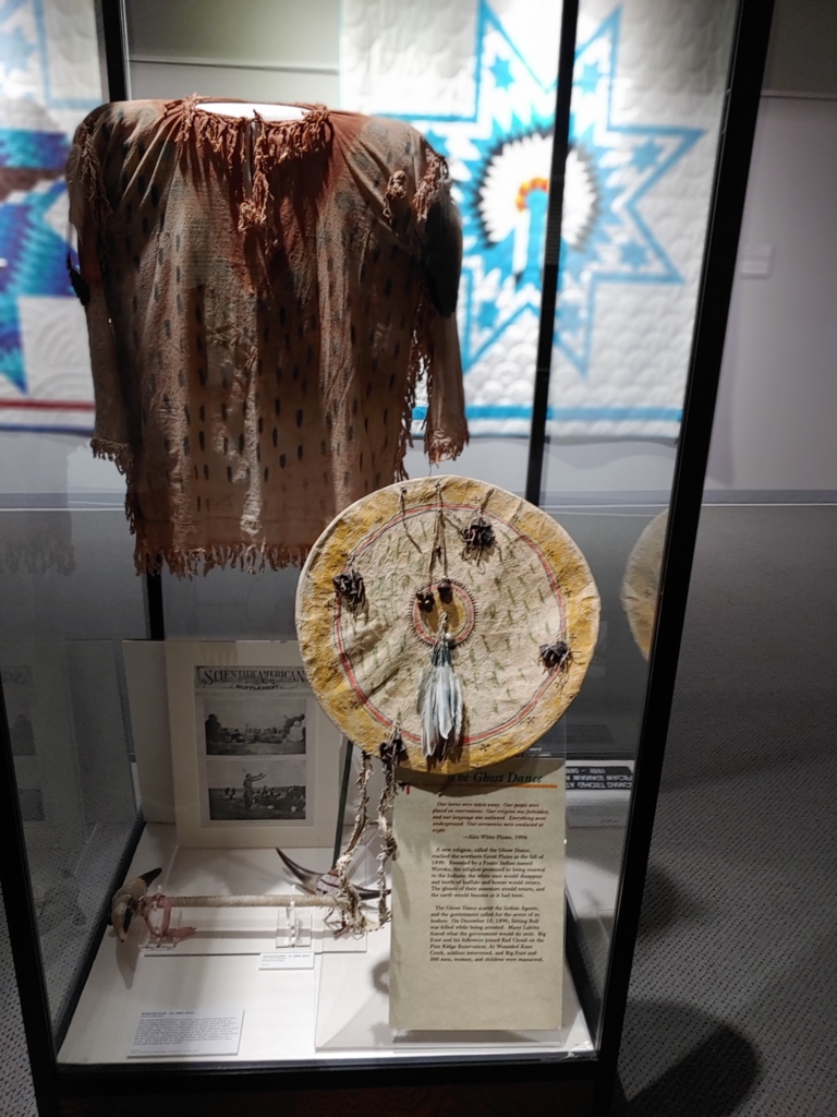 Photo of a museum display featuring Ghost dance regaila and drum, as well as photos and a text description that is illegible in the photograph.