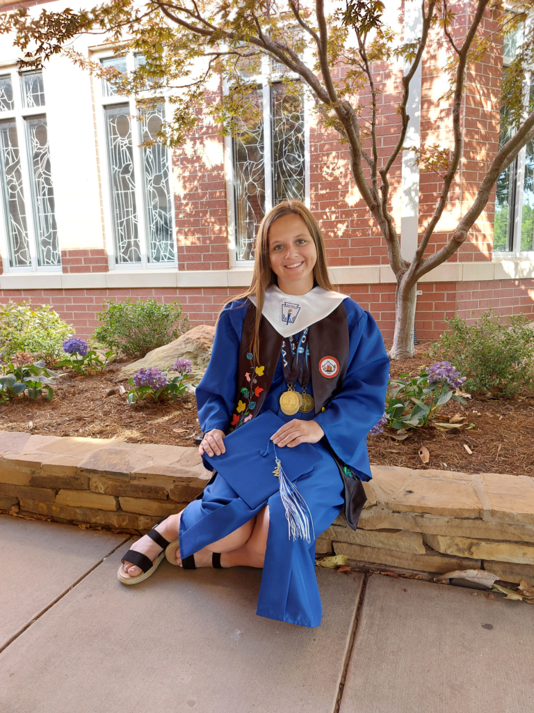Tribal member Shelby O'Steen sits on a ledge in front of a brick wall. She wears a bright blue high school graduation gown and a CPN graduation stole, and holds her graduation cap in her hands. 