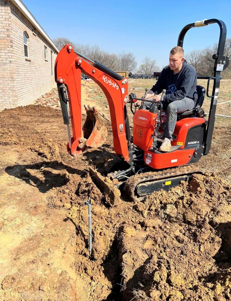 A man with short, light hair wearing jeans and a long-sleeved shirt uses an orange excavator to dig a hole.
