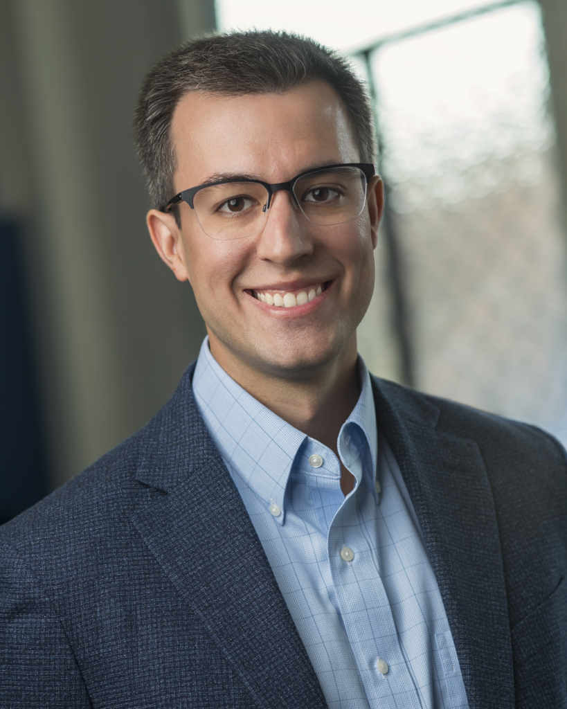 Headshot of CPN tribal member Andrew Mock. Wears glasses with dark upper rims, and a navy suit and light blue check shirt.