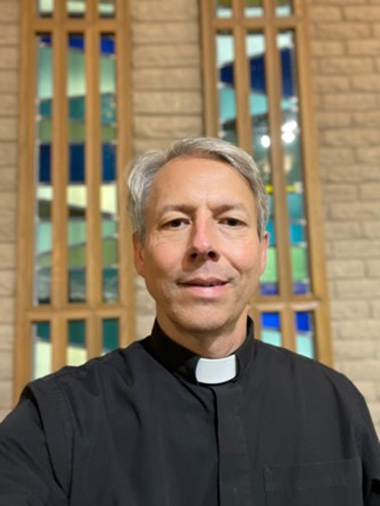 Portrait of Rev. John Henson in a priest's collar and attire in front of a stained glass window.