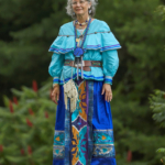 Full-body portrait of photographer and CPN Tribal member Sharon Hoogstraten in regalia of dark and teal blues with orange floral accents. She stands on a rock in front of some trees, and the light suggests a slight cloud cover.