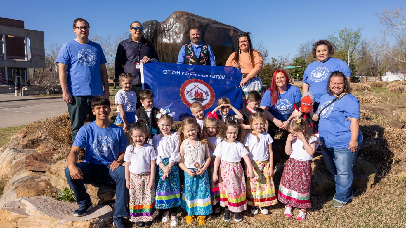 A group of young children wearing ribbon skirts and vests stand with several adult language instructors wearing blue shirts with the Citizen Potawatomi Nation seal on them and holding a CPN flag outside of the Same Noble History Museum near a bison statue.