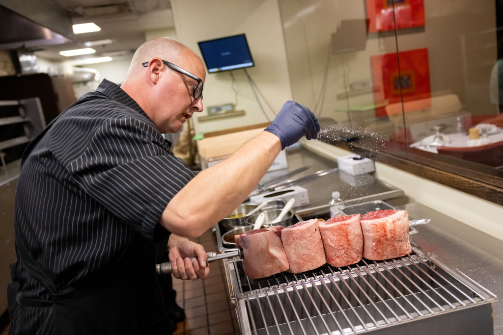 A chef with a bald head, dark glasses, a black shirt with white pinstripes, and wearing rubber gloves tosses salt over four large pieces of meat on a skewer.