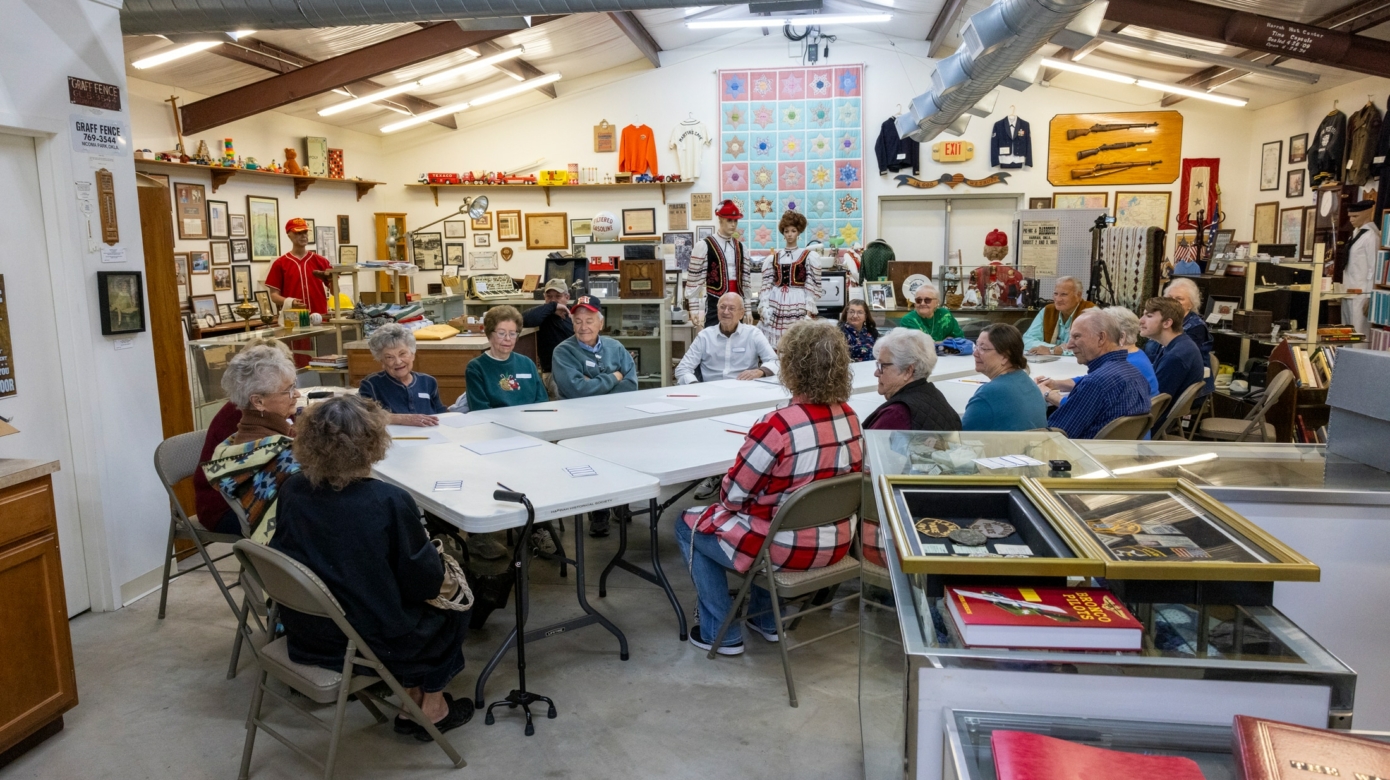 Tribal members, mostly elders, sig around white folding tables in a museum storeroom full of mannequins, picture frames, and other objects.