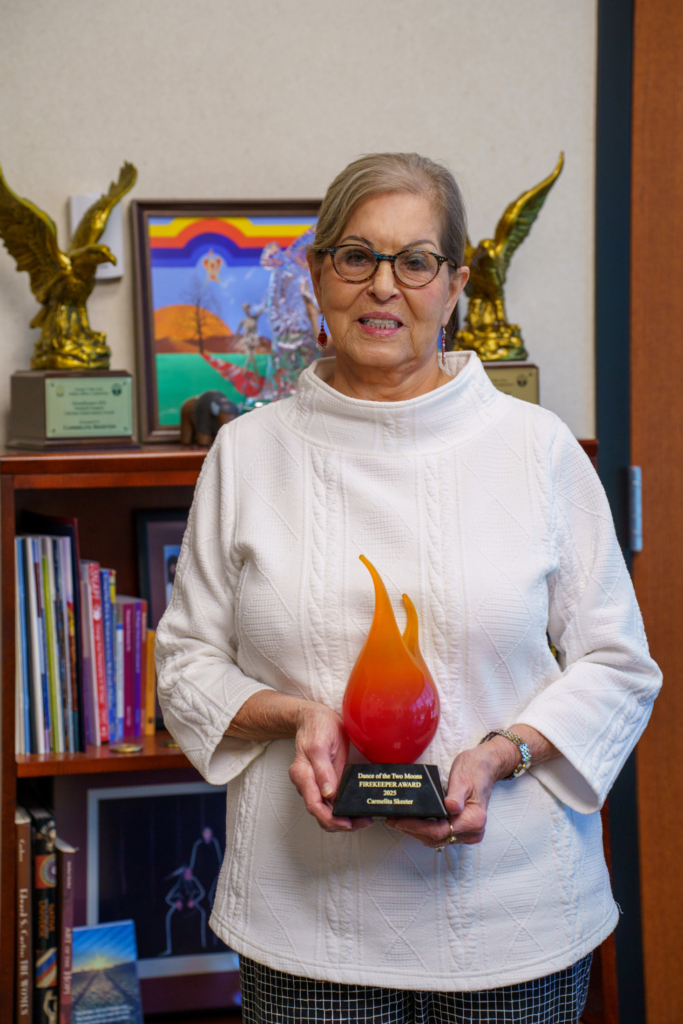 A person in a white turtleneck sweater, black glasses, and gray hair pulled back behind their head holds an award shaped like a flame.