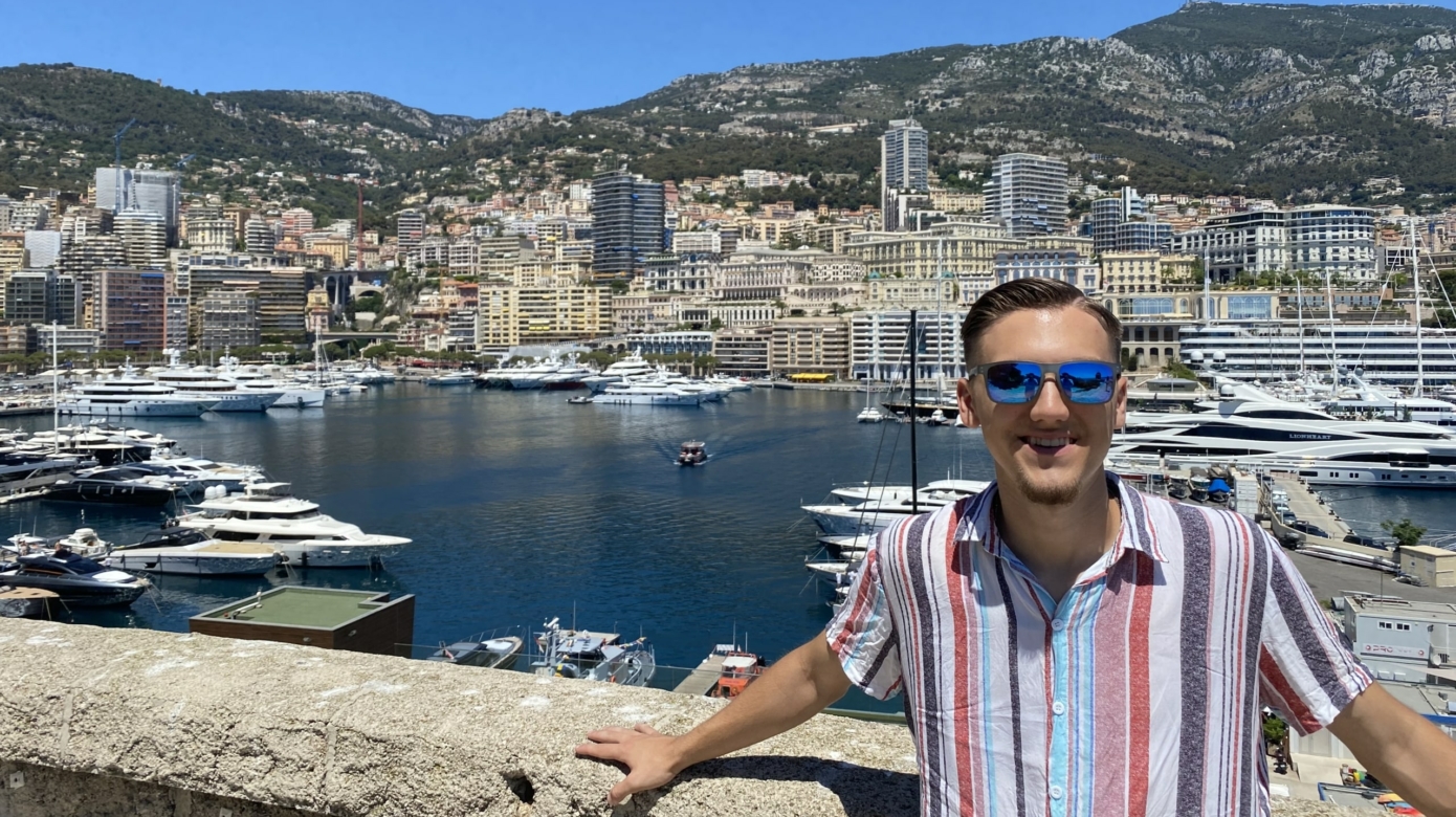 CPN tribal member Jake Biddy stands in front of the water front on the French Riviera. He wears a pink and blue vertical striped shirt and sunglasses, and leans against a bridge.