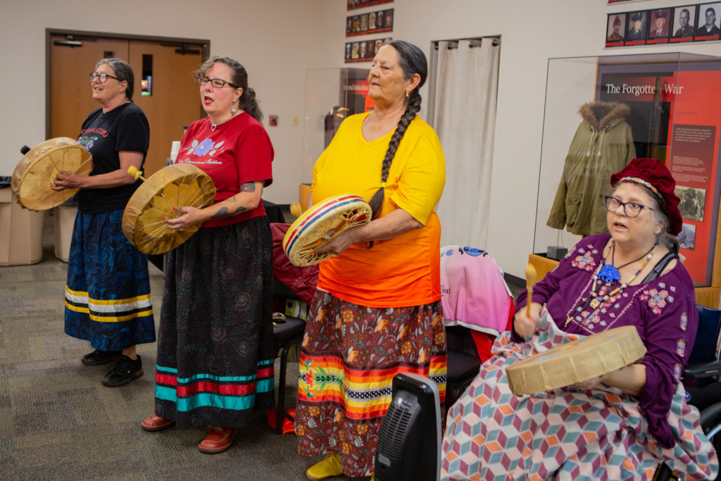 Four people in bright colored ribbon skirts play hand drums and sing.