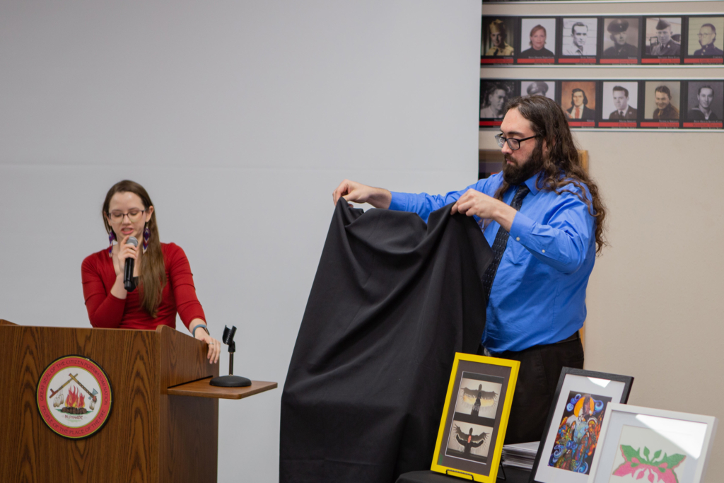 A person wearing a blue shirt and black tie lifts a black cloth from an easel. A person in red speaks into a microphone at a podium.