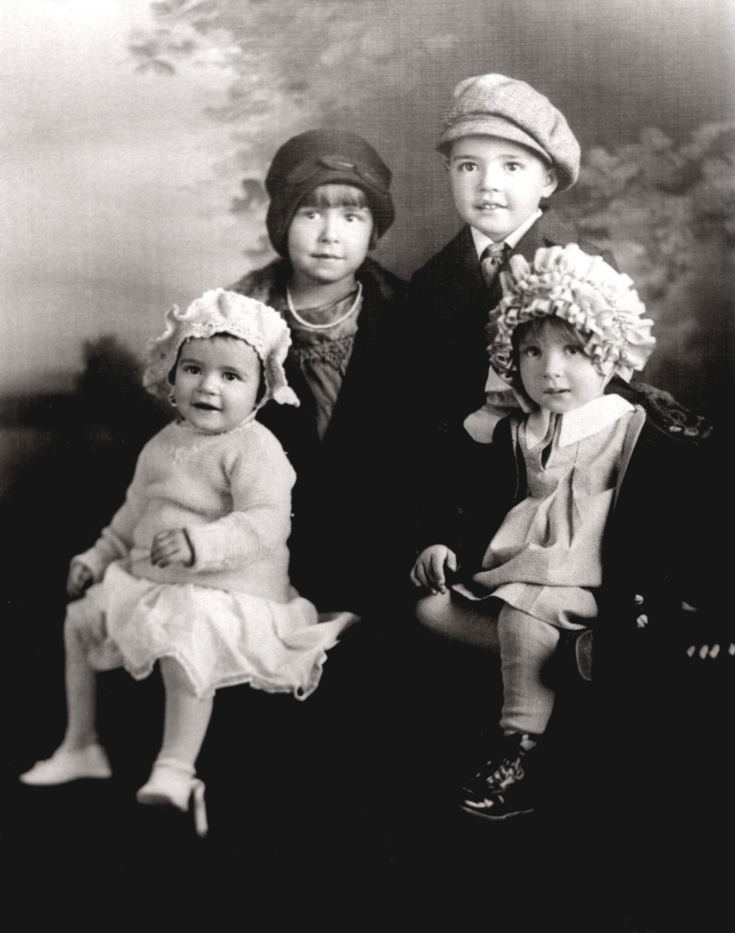 Black and white photograph of four children, the two oldest standing and the two youngest seated in front of them.