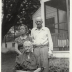 Archival photo of Julia McEvers, Julia Anderson, and Dave McEvers. Julia McEvers and Dave McEvers stand behind a seated Julia Anderson in the front yard of a house.