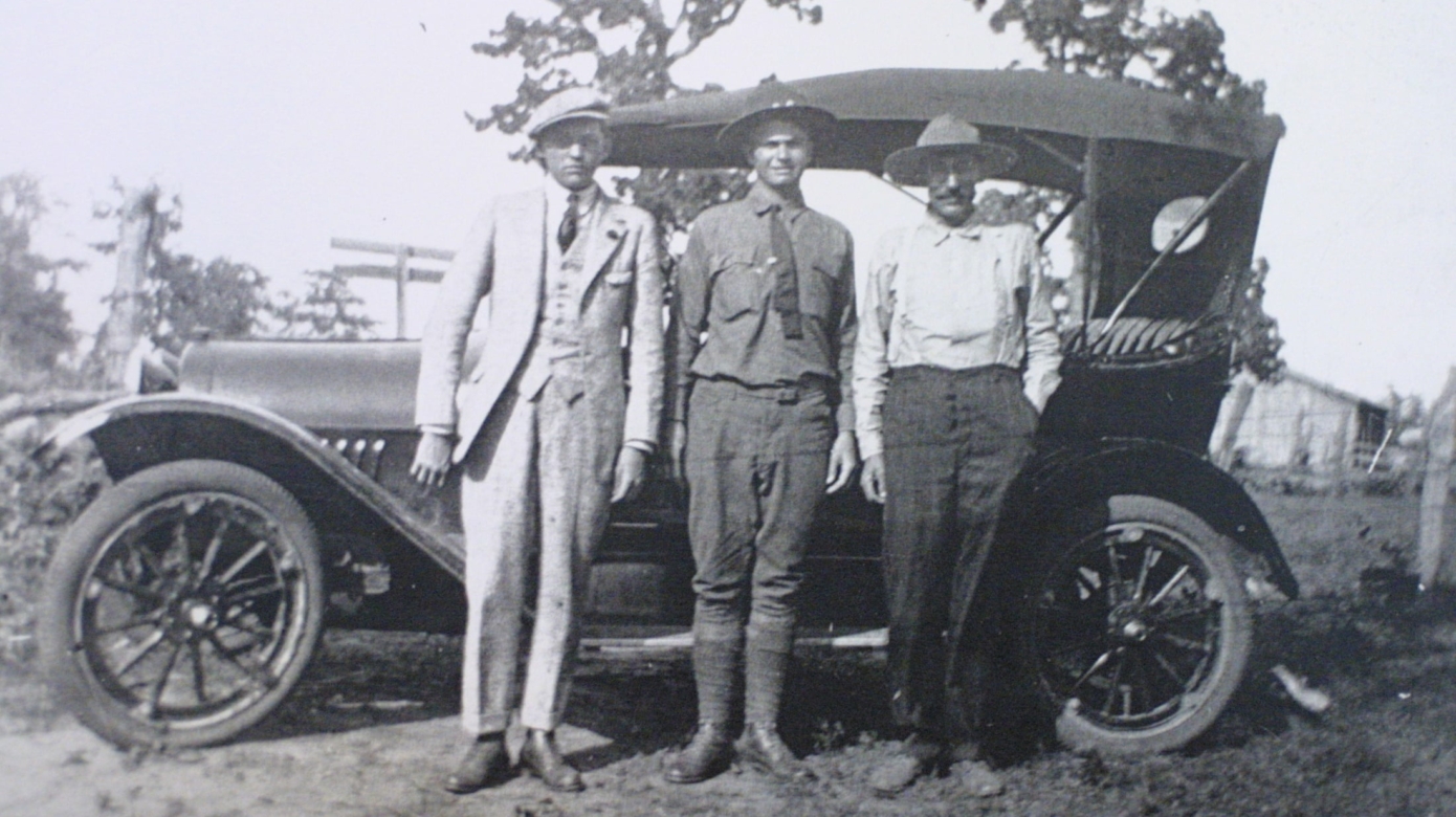 Black and white photo of three men in business wear and hats stand in front of an early 20th century car.