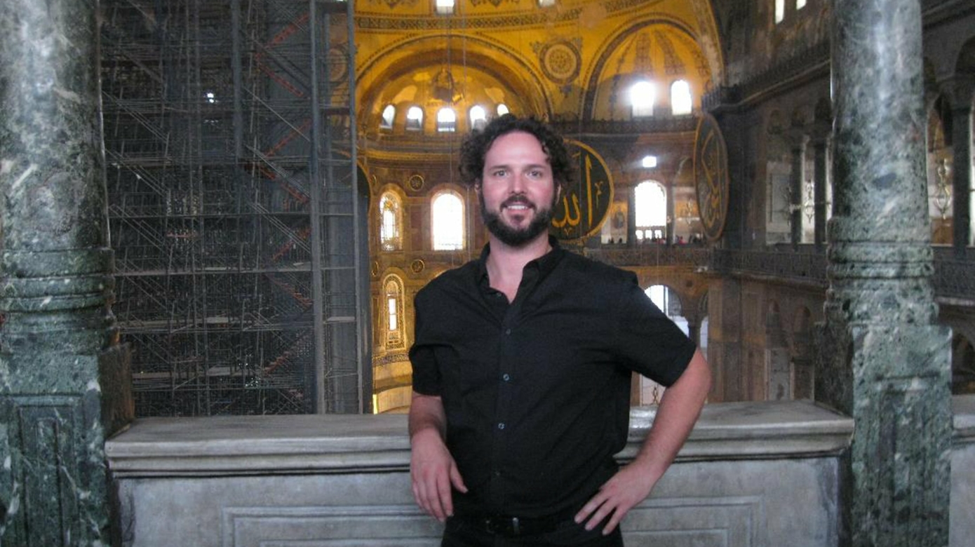 Tribal member Preston Stovall poses in the Holy Hagia Sophia Grand Mosque in Istanbul, Turkey. He wears all black, and dark hair and beard frame his face. Behind him are a large expanse of a stone hall, filled with arched windows. In the far background, light reveals the colors of the patterning on the walls and domes.